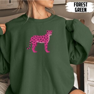 May include: Forest green sweatshirt featuring a pink leopard illustration with black spots. The text "FOREST GREEN" is printed in white on the upper right. The sweatshirt is a solid color with a crew neck.