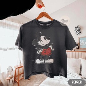 Może przedstawiać: Ciemnoszara koszulka z nadrukiem Mickey Mouse w stylu vintage w kolorach czarnym, białym i czerwonym. Koszulka wisi na drewnianym wieszaku. Na dole obrazu widnieje napis "Pepper".