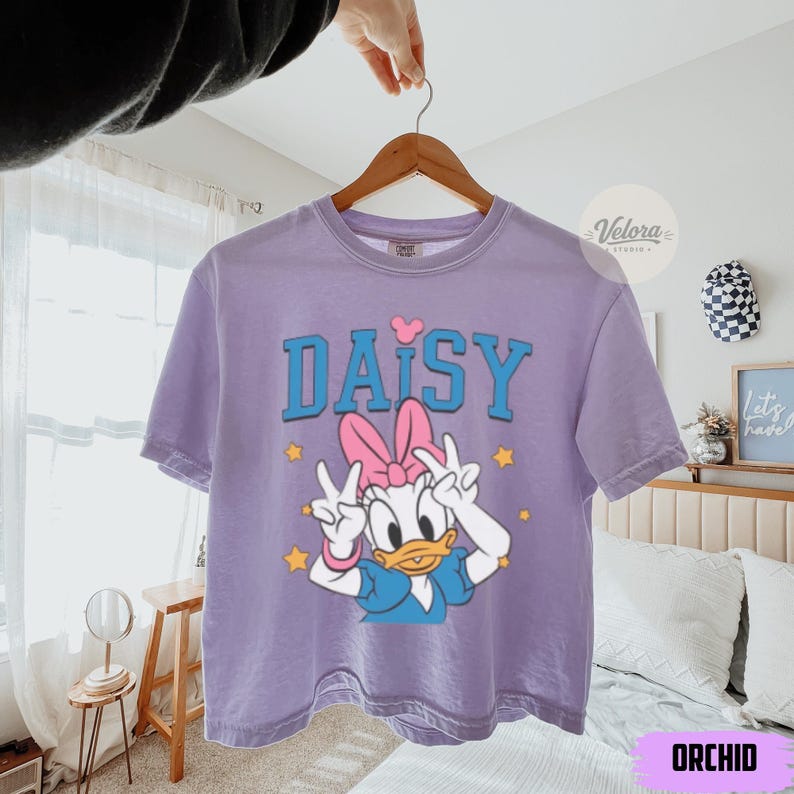 May include: A lilac t-shirt featuring the word "DAISY" in blue block letters and a pink heart above the "I". The shirt has a cartoon Daisy Duck graphic with a pink bow and peace signs. Yellow stars are scattered around the graphic.