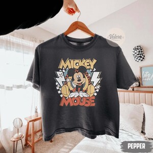 Puede incluir: Una camiseta gris oscuro con un dise&ntilde;o de Mickey Mouse. El dise&ntilde;o incluye las palabras "Mickey Mouse" en amarillo y rojo, con Mickey sentado. La camiseta est&aacute; colgada en una percha de madera.