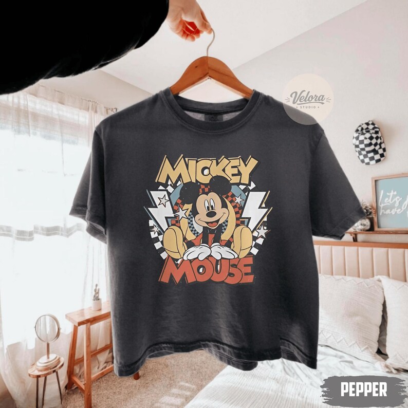 Puede incluir: Una camiseta gris oscuro con un dise&ntilde;o de Mickey Mouse. El dise&ntilde;o incluye las palabras "Mickey Mouse" en amarillo y rojo, con Mickey sentado. La camiseta est&aacute; colgada en una percha de madera.
