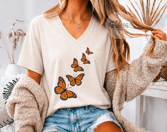 May include: A cream-colored V-neck t-shirt featuring a graphic print of five orange and black monarch butterflies arranged in a diagonal upward pattern. The shirt is worn with a textured beige cardigan and light blue denim shorts.