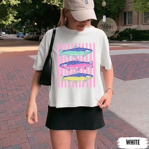 May include: White t-shirt featuring three colorful fish designs on a pink striped background. The fish are in shades of blue, pink, and yellow. The word "WHITE" is at the bottom of the image.