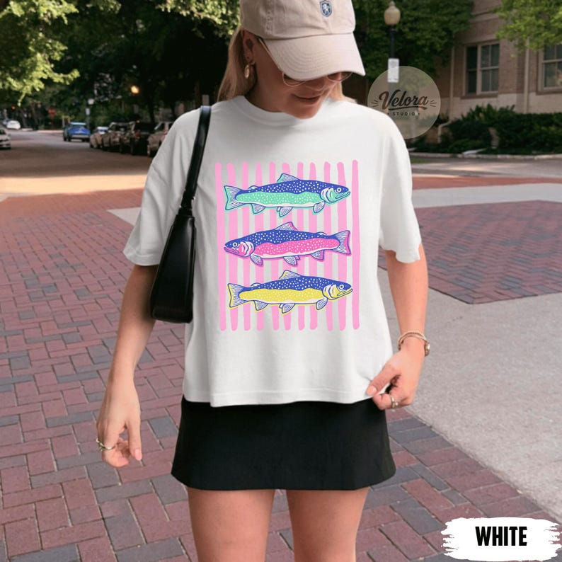 May include: White t-shirt featuring three colorful fish designs on a pink striped background. The fish are in shades of blue, pink, and yellow. The word "WHITE" is at the bottom of the image.