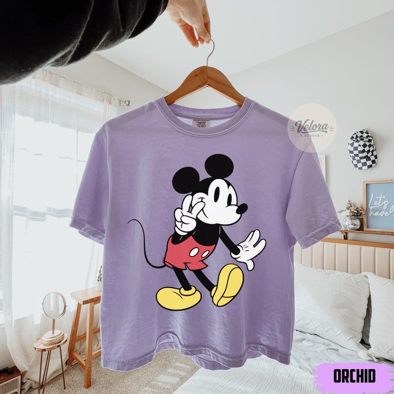 May include: A lavender t-shirt featuring a graphic of Mickey Mouse in a classic pose. Mickey is depicted with black ears, a white face, red shorts, yellow shoes, and white gloves. The shirt is hanging on a wooden hanger.