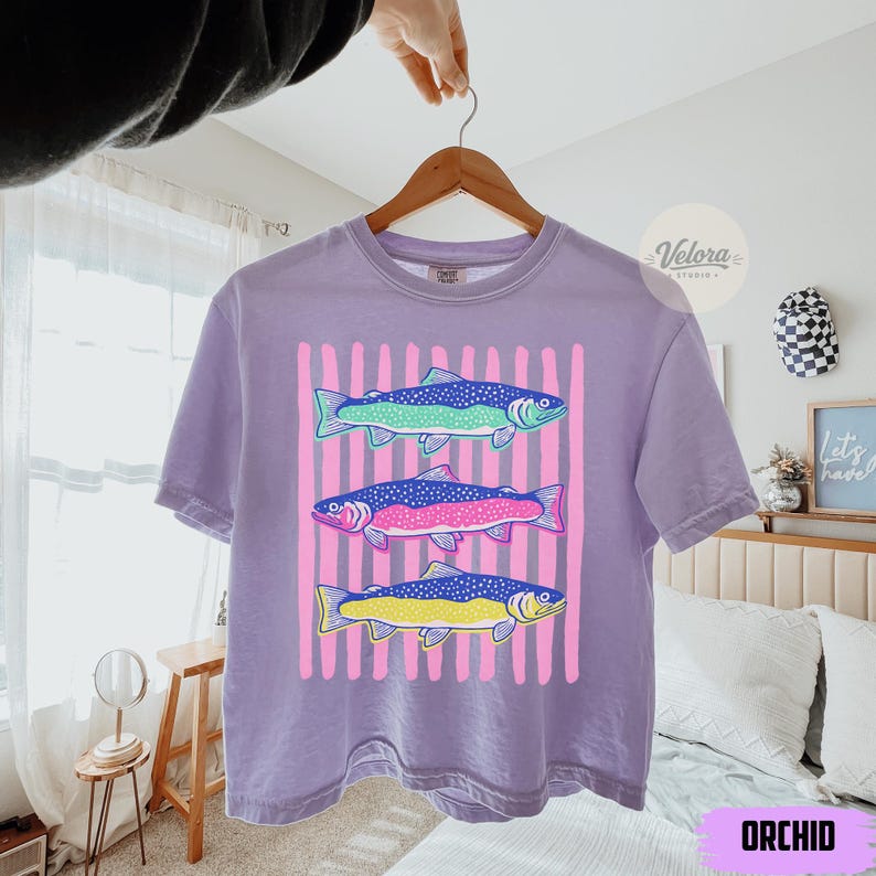 May include: Orchid purple t-shirt featuring a colorful fish design. The graphic includes three fish in shades of blue, pink, and yellow, set against a backdrop of pink vertical stripes. The shirt is hanging on a wooden hanger.