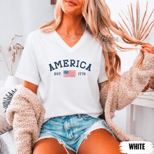 May include: White V-neck t-shirt with the word "AMERICA" in navy blue, the American flag, and the year "1776". The shirt is a casual style.