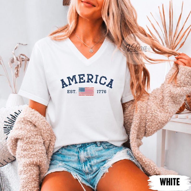 May include: White V-neck t-shirt with the word "AMERICA" in navy blue, the American flag, and the year "1776". The shirt is a casual style.