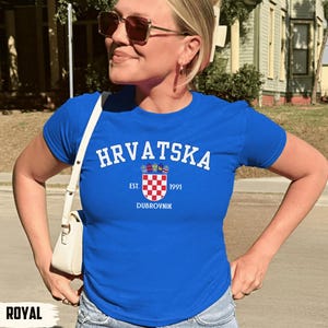 May include: Royal blue t-shirt with the word "HRVATSKA" in white arched text. Below the text is a crest with a checkered pattern and the text "EST. 1991 DUBROVNIK". The shirt is short-sleeved.