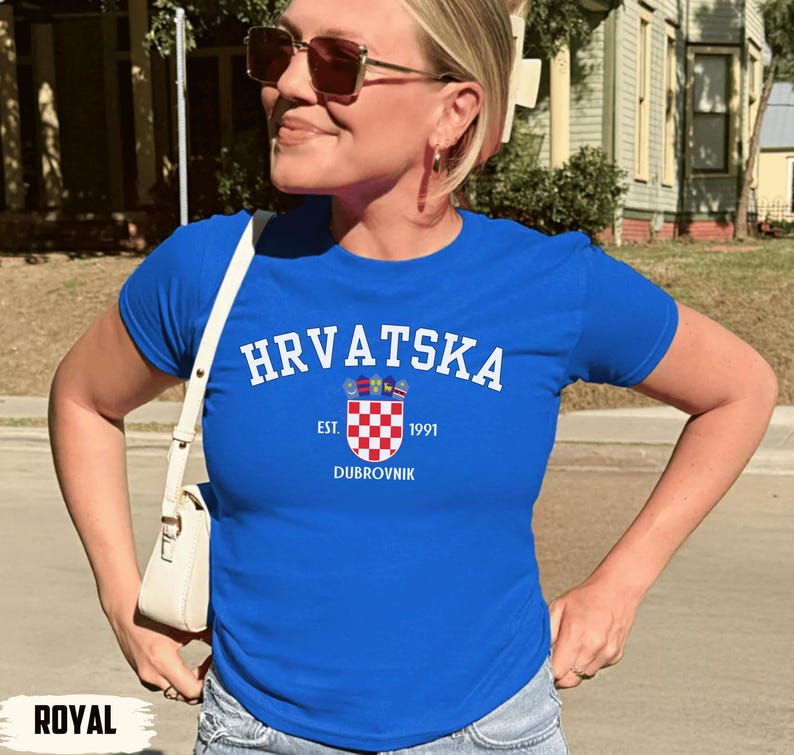 May include: Royal blue t-shirt with the word "HRVATSKA" in white arched text. Below the text is a crest with a checkered pattern and the text "EST. 1991 DUBROVNIK". The shirt is short-sleeved.
