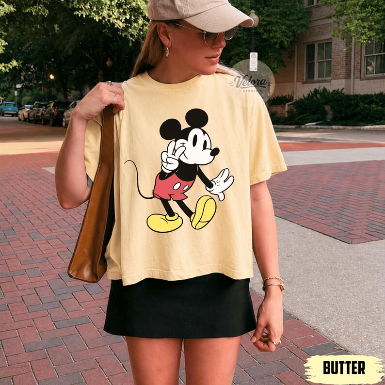May include: Butter-colored t-shirt featuring a graphic of Mickey Mouse giving a peace sign. Mickey is depicted in his classic outfit with red shorts, yellow shoes, and white gloves. The shirt is paired with a black skirt.