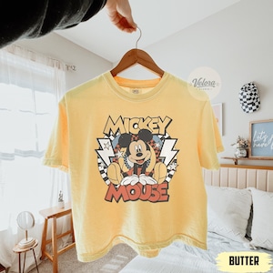 Puede incluir: Camiseta amarilla mantequilla con un gr&aacute;fico de Mickey Mouse. El dise&ntilde;o incluye las palabras "Mickey Mouse" en una fuente retro, con Mickey en el centro. La camiseta est&aacute; colgada en una percha de madera.