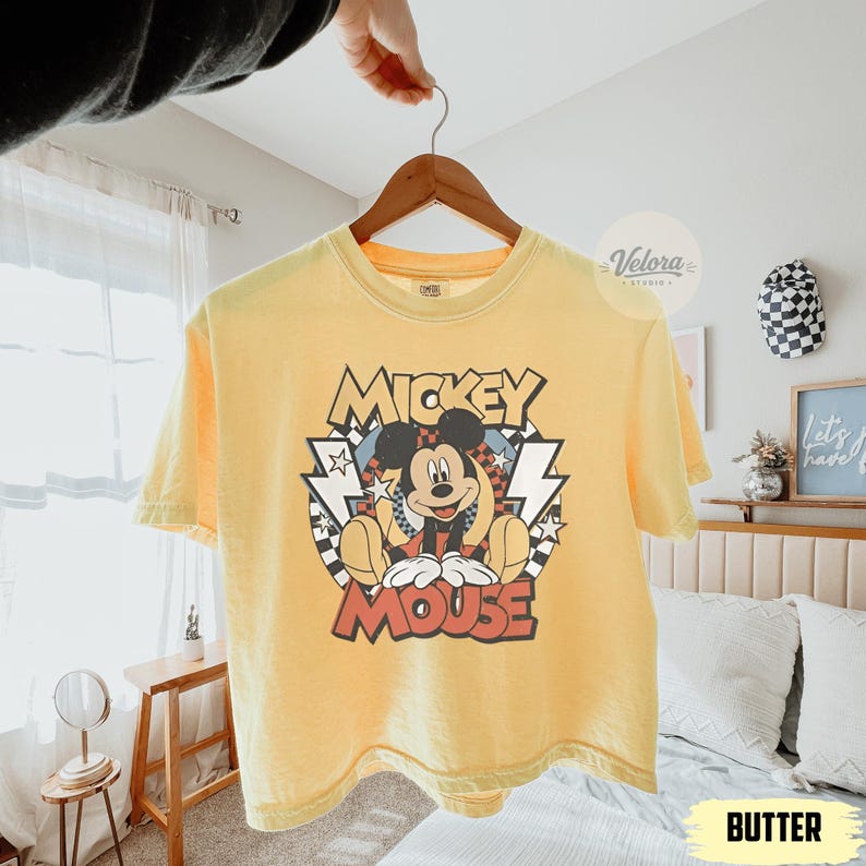Puede incluir: Camiseta amarilla mantequilla con un gr&aacute;fico de Mickey Mouse. El dise&ntilde;o incluye las palabras "Mickey Mouse" en una fuente retro, con Mickey en el centro. La camiseta est&aacute; colgada en una percha de madera.