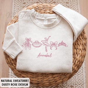 Puede incluir: Una sudadera de color beige claro con un dise&ntilde;o rosa empolvado que presenta zapatillas de ballet, un tut&uacute;, una bailarina, lazos y una corona. El nombre "Annabel" est&aacute; escrito en cursiva debajo. El texto "NATURAL SWEATSHIRT DUSTY ROSE DESIGN" est&aacute; en la parte inferior.