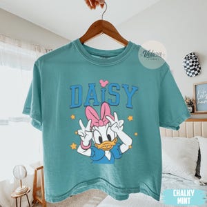 May include: A teal t-shirt with a Daisy Duck graphic, giving a peace sign. The word "DAISY" is printed in blue block letters above the character. A pink bow and stars are also featured in the design. The shirt is hanging on a wooden hanger.