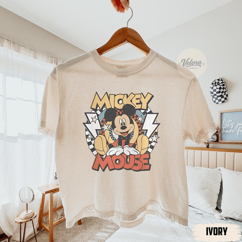 Puede incluir: Camiseta de color marfil con un dise&ntilde;o de Mickey Mouse. El dise&ntilde;o incluye las palabras "Mickey Mouse" en una fuente retro, con Mickey en el centro. La camiseta est&aacute; colgada en una percha de madera.