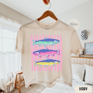 May include: Ivory-colored t-shirt featuring a graphic of three colorful fish. The fish are blue, green, pink, and yellow, set against a pink striped background. The shirt is hanging on a wooden hanger.