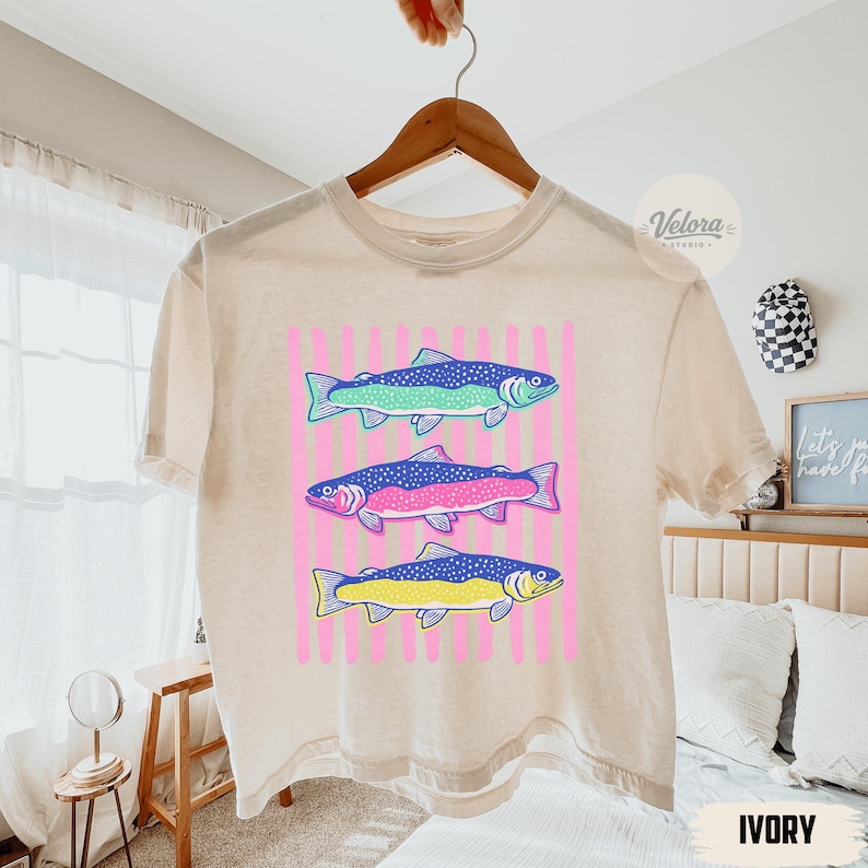 May include: Ivory-colored t-shirt featuring a graphic of three colorful fish. The fish are blue, green, pink, and yellow, set against a pink striped background. The shirt is hanging on a wooden hanger.