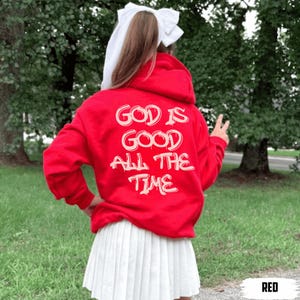 May include: A red hoodie with the text "GOD IS GOOD ALL THE TIME" in white lettering. The hoodie is paired with a white pleated skirt. The image is taken outdoors with a green background.