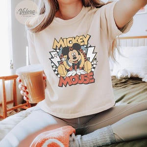 Puede incluir: Camiseta de color marfil con un dise&ntilde;o retro de Mickey Mouse y las palabras "Mickey Mouse". El conjunto incluye leggings grises y zapatillas naranjas. Se sostiene en la mano un vaso de pl&aacute;stico transparente con una bebida marr&oacute;n.