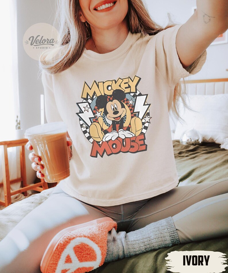 Puede incluir: Camiseta de color marfil con un dise&ntilde;o retro de Mickey Mouse y las palabras "Mickey Mouse". El conjunto incluye leggings grises y zapatillas naranjas. Se sostiene en la mano un vaso de pl&aacute;stico transparente con una bebida marr&oacute;n.