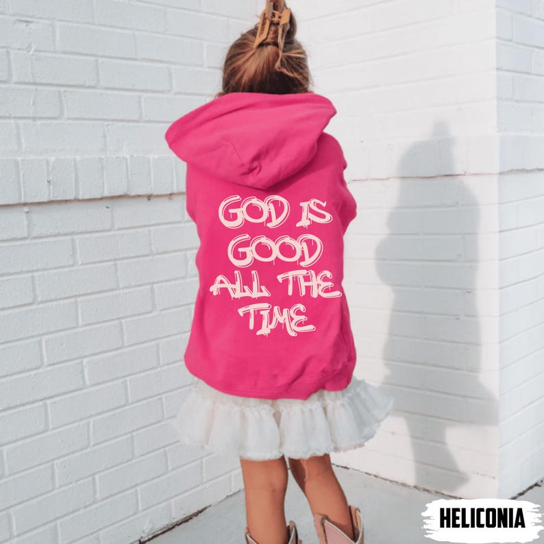 May include: A hot pink hooded sweatshirt with the text "GOD IS GOOD ALL THE TIME" in white. The sweatshirt is worn by a person, paired with a white ruffled skirt and cowboy boots. The background is a white brick wall.