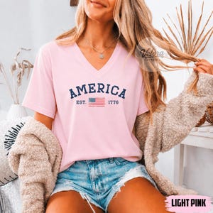 May include: A light pink V-neck t-shirt with the word "AMERICA" in navy blue, along with "EST. 1776" and a small American flag graphic. The shirt is a short-sleeved top.