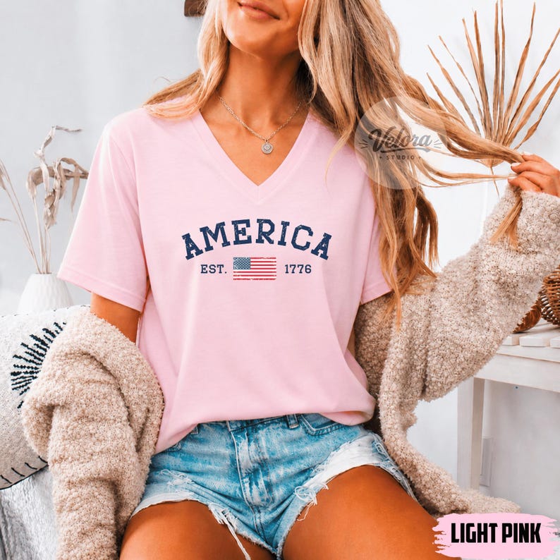 May include: A light pink V-neck t-shirt with the word "AMERICA" in navy blue, along with "EST. 1776" and a small American flag graphic. The shirt is a short-sleeved top.