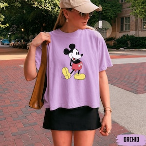 May include: A light purple t-shirt featuring a classic Mickey Mouse graphic. The cartoon character is depicted in black, red, yellow, and white. The word "ORCHID" is printed at the bottom of the image.