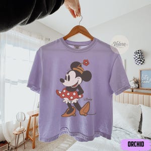 May include: Orchid-colored t-shirt featuring a Minnie Mouse graphic. The cartoon character is depicted in a classic pose, wearing a red polka-dot skirt, a hat with a flower, and brown shoes. The word "Disney" is printed below the character.