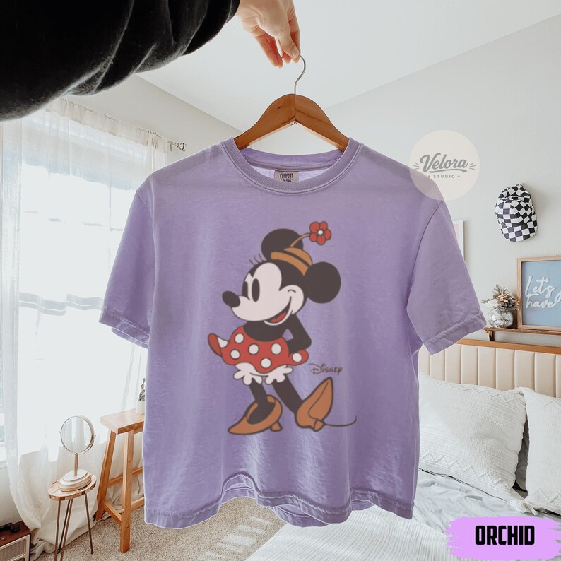 May include: Orchid-colored t-shirt featuring a Minnie Mouse graphic. The cartoon character is depicted in a classic pose, wearing a red polka-dot skirt, a hat with a flower, and brown shoes. The word "Disney" is printed below the character.