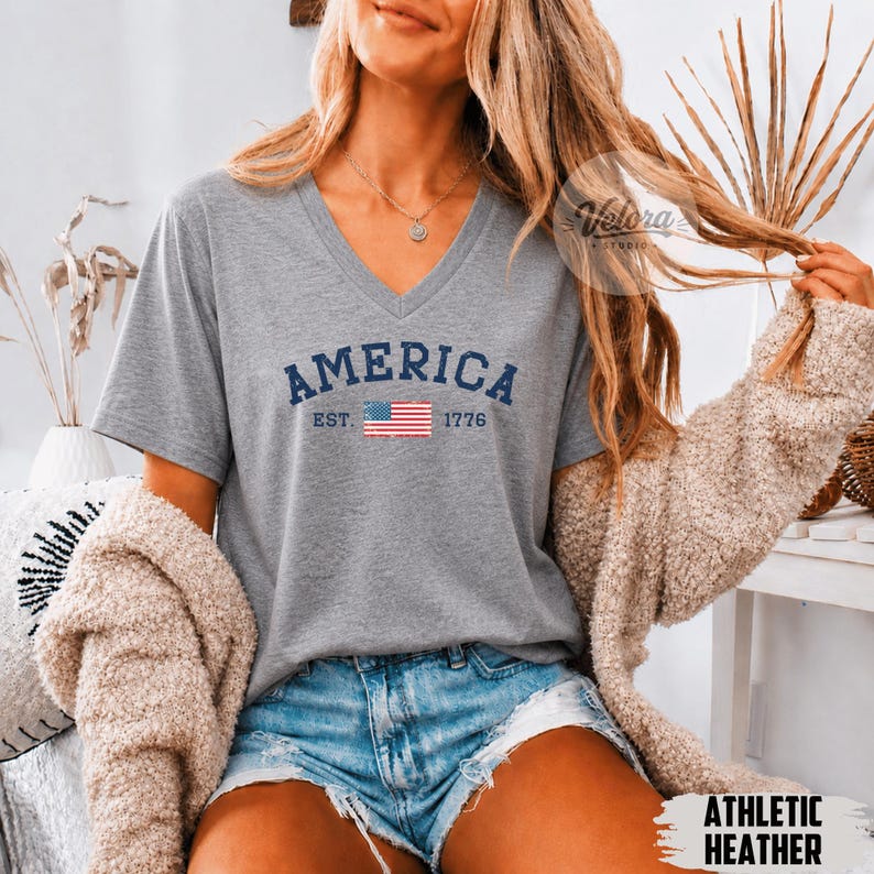 May include: A heather gray V-neck t-shirt with the word "AMERICA" in navy blue, the American flag, and the text "EST. 1776". The shirt is made of a soft material and is suitable for casual wear.