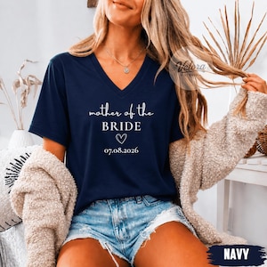 May include: A navy blue V-neck t-shirt featuring white text that reads mother of the BRIDE, a small heart outline, and the date 07.08.2026. The shirt is worn with a beige textured cardigan and light blue denim shorts with a frayed hemline.