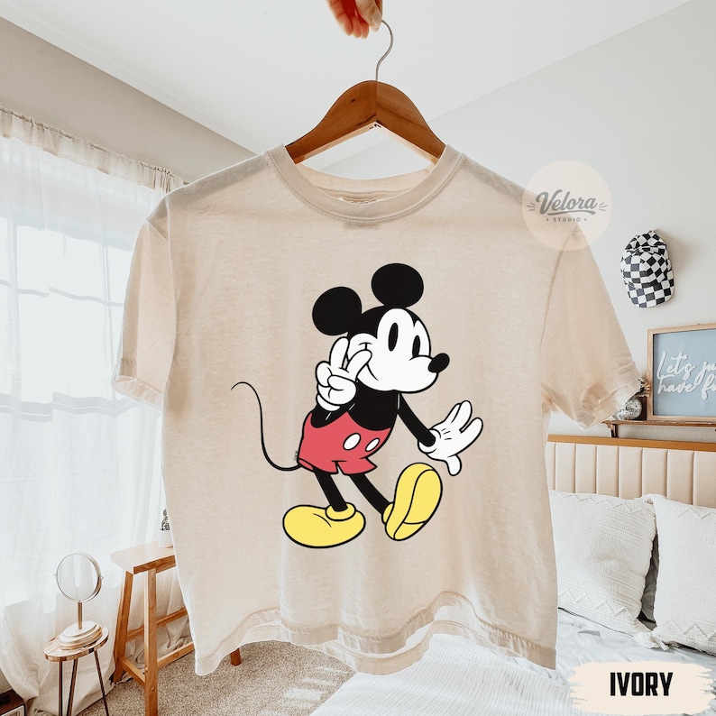 May include: Ivory-colored t-shirt featuring a classic Mickey Mouse graphic. The cartoon character is depicted in black, red, yellow, and white, with a peace sign gesture. The shirt is hanging on a wooden hanger.