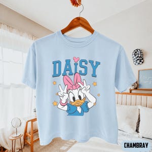 May include: Pale blue t-shirt with a Daisy Duck graphic. The design includes a pink bow, peace signs, and the word "DAISY" in blue. Yellow stars are also present. The t-shirt is displayed on a wooden hanger.