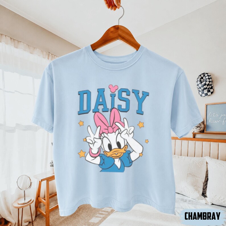 May include: Pale blue t-shirt with a Daisy Duck graphic. The design includes a pink bow, peace signs, and the word "DAISY" in blue. Yellow stars are also present. The t-shirt is displayed on a wooden hanger.