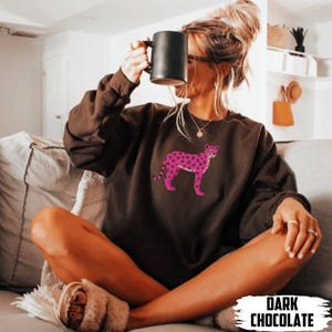 May include: Dark chocolate-colored sweatshirt featuring a pink leopard design. The sweatshirt has a crew neck and long sleeves. The text "DARK CHOCOLATE" is at the bottom of the image. The sweatshirt is worn by a person.
