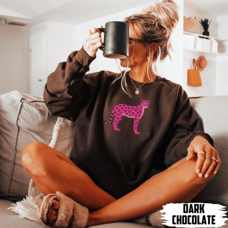 May include: Dark chocolate-colored sweatshirt featuring a pink leopard design. The sweatshirt has a crew neck and long sleeves. The text "DARK CHOCOLATE" is at the bottom of the image. The sweatshirt is worn by a person.