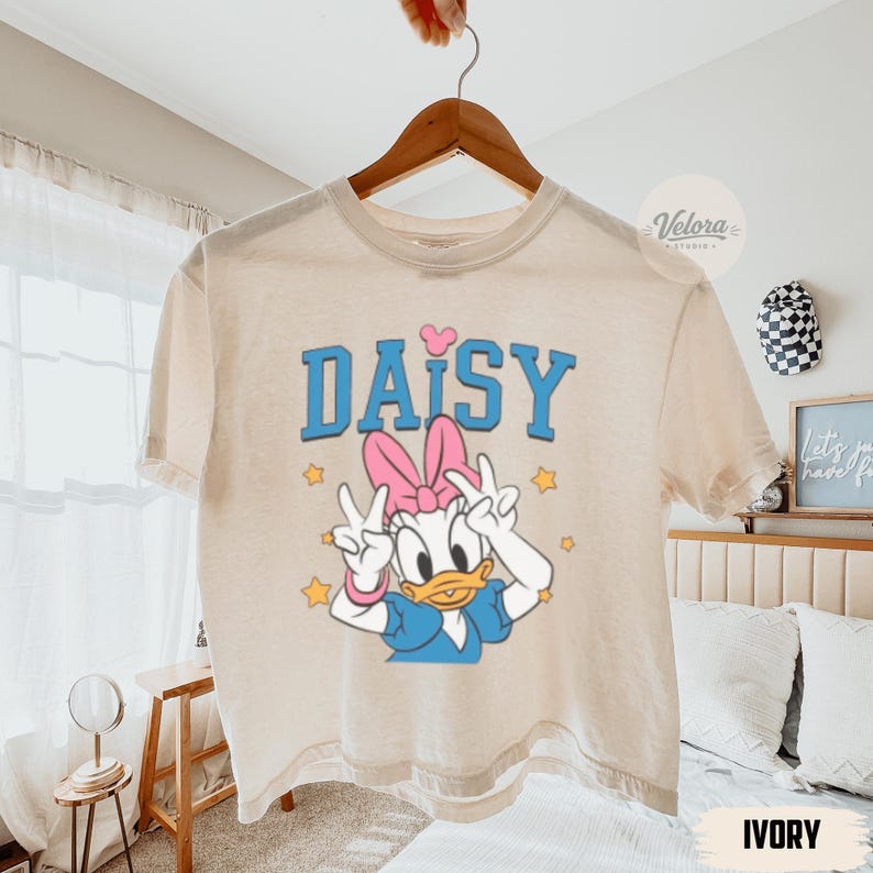 May include: Ivory t-shirt with a Daisy Duck graphic, featuring a pink bow, blue top, and peace signs. The word "DAISY" is printed in blue block letters with a pink heart above the "I". Gold stars surround the character. The shirt is displayed on a wooden hanger.