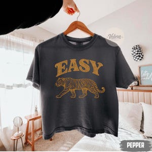 Puede incluir: Una camiseta gris oscuro con la palabra "EASY" en letras doradas sobre una ilustración de un tigre dorado. La camiseta está colgada de una percha de madera.