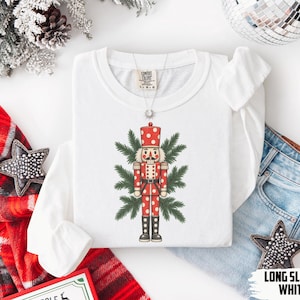 May include: A white long-sleeve shirt featuring a graphic of a red and white polka-dot nutcracker centered in front of green pine branches. The shirt has a crew neckline and a tag at the collar that reads Comfort Colors with size L-G-G.