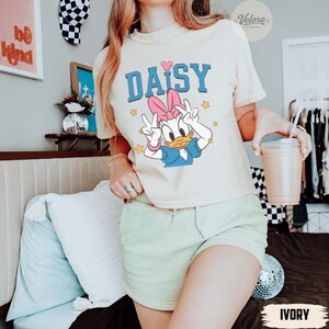 May include: Ivory t-shirt with a Daisy Duck graphic and the word "DAISY" in blue. Daisy is giving a peace sign. The shirt is worn with light green shorts. A clear plastic cup with a straw is held in the hand. The word "IVORY" is at the bottom.