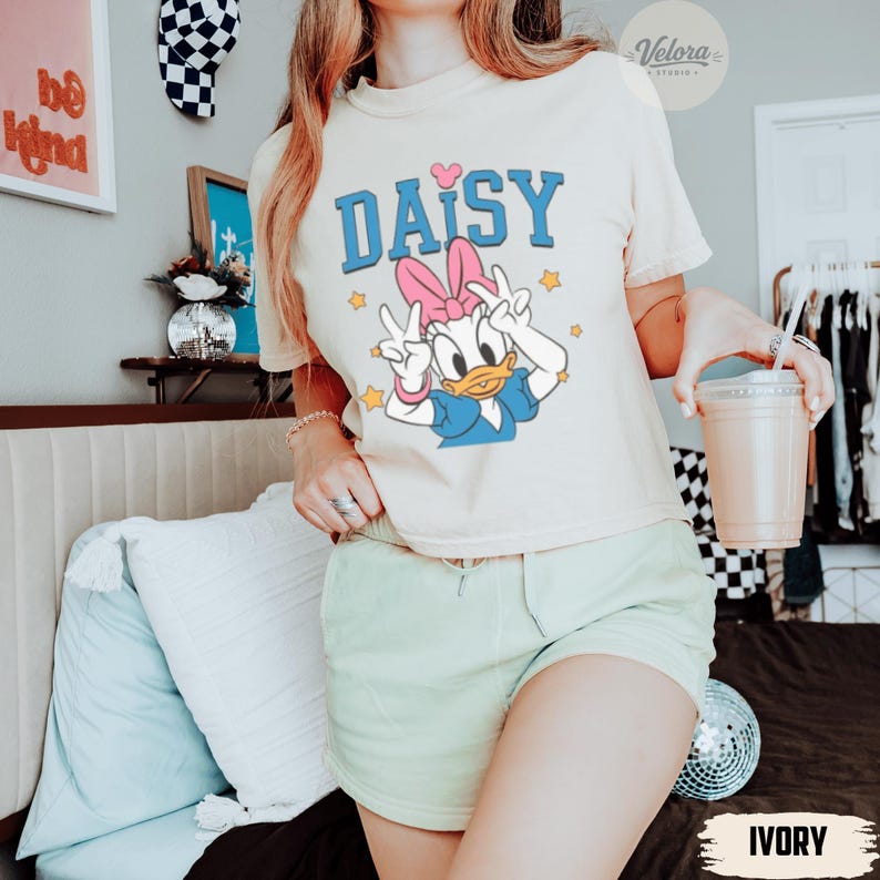 May include: Ivory t-shirt with a Daisy Duck graphic and the word "DAISY" in blue. Daisy is giving a peace sign. The shirt is worn with light green shorts. A clear plastic cup with a straw is held in the hand. The word "IVORY" is at the bottom.