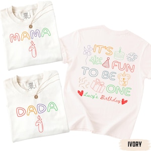 Op de afbeelding: Ivoor kleurige t-shirts met kleurrijke tekst en afbeeldingen. Eén shirt zegt "MAMA" met een kaarsafbeelding, een ander zegt "DADA" met een kaars, en een derde zegt "IT'S FUN TO BE ONE" met verjaardagsillustraties en "Lucy's Birthday".