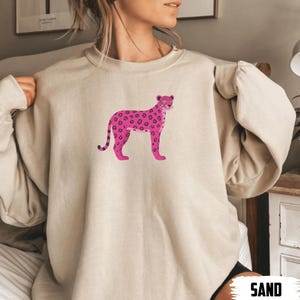 May include: A sand-colored sweatshirt featuring a pink leopard design. The leopard has dark pink spots and is standing in a side profile. The word "SAND" is printed in white at the bottom right corner of the image.