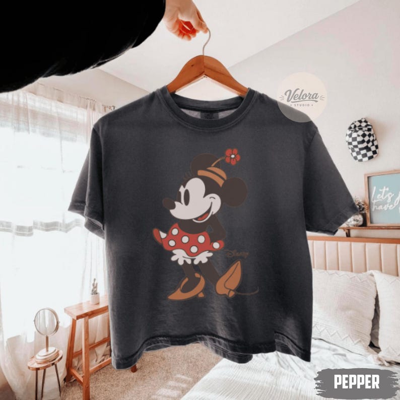 May include: A dark gray t-shirt featuring a cartoon Minnie Mouse design. Minnie is depicted in a red polka-dot dress, a matching bow, and brown shoes. The shirt is hanging on a wooden hanger.