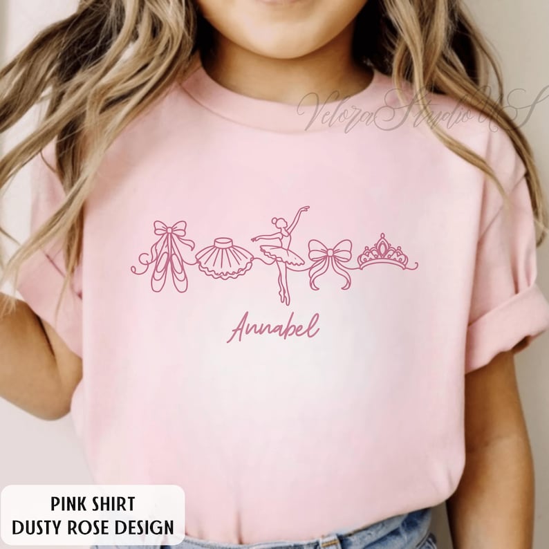 Puede incluir: Una camiseta rosa con un dise&ntilde;o rosa polvoriento con ilustraciones tem&aacute;ticas de ballet. El dise&ntilde;o incluye zapatillas de ballet, un tut&uacute;, una bailarina, un lazo y una corona. El nombre "Annabell" est&aacute; escrito en cursiva.
