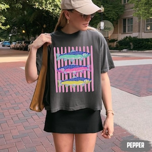 May include: Dark gray t-shirt featuring three colorful fish designs on a pink striped background. The fish are in shades of blue, pink, and yellow. The word "PEPPER" is printed at the bottom of the image.
