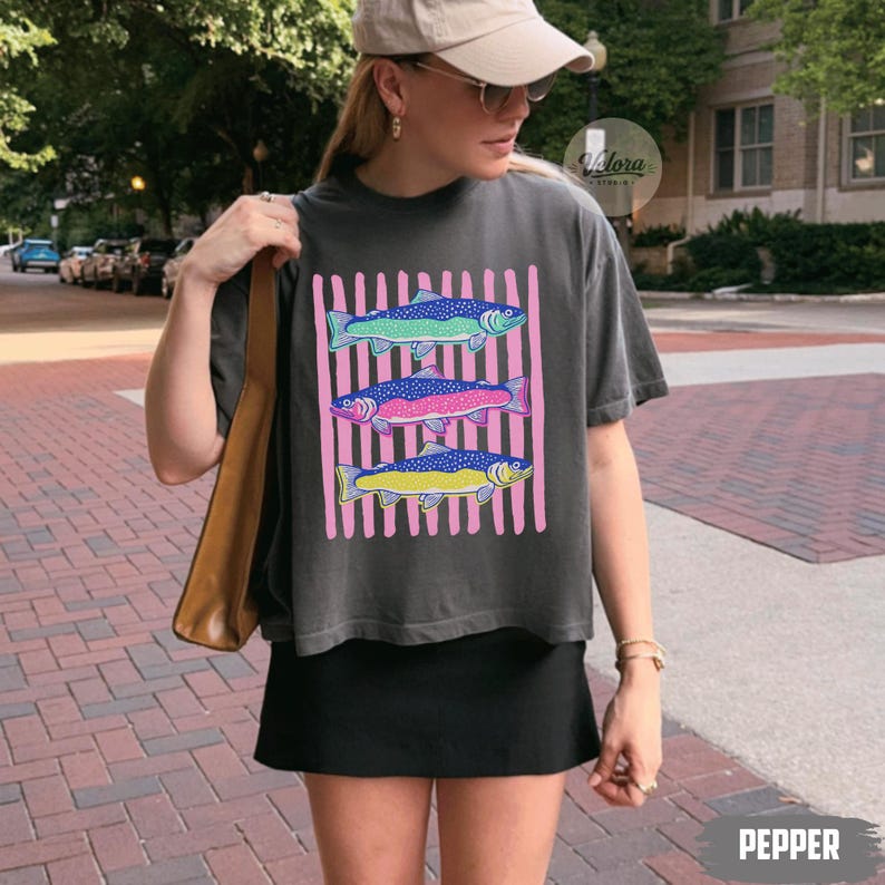May include: Dark gray t-shirt featuring three colorful fish designs on a pink striped background. The fish are in shades of blue, pink, and yellow. The word "PEPPER" is printed at the bottom of the image.