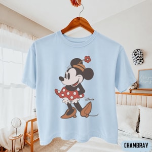 May include: Light blue t-shirt featuring a vintage-style illustration of Minnie Mouse. Minnie is depicted in a red polka-dot dress, brown shoes, and a hat with a flower. The Disney logo is visible. The shirt is hanging on a wooden hanger.